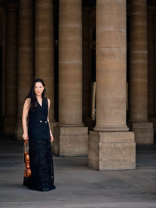 Anna Lee standing with her violin in a long stone colonnade