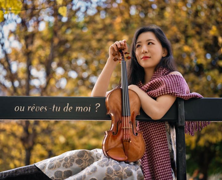 Anna Lee seated on a park bench with her violin among autumn leaves