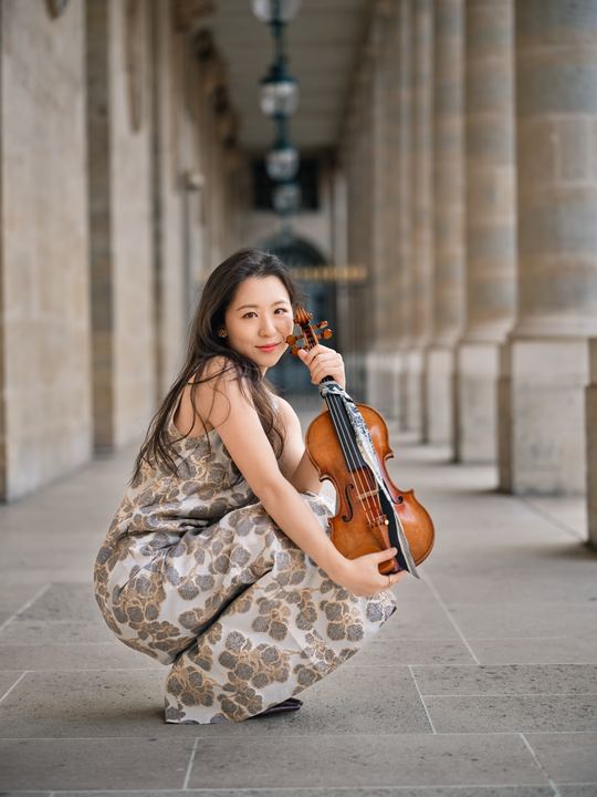 Anna Lee with her violin in a patterned gown in a colonnade