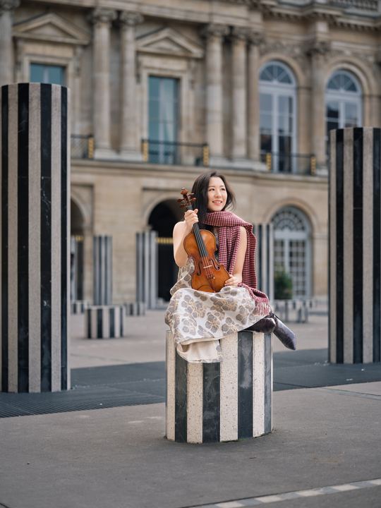Anna Lee seated with her violin among black-and-white columns